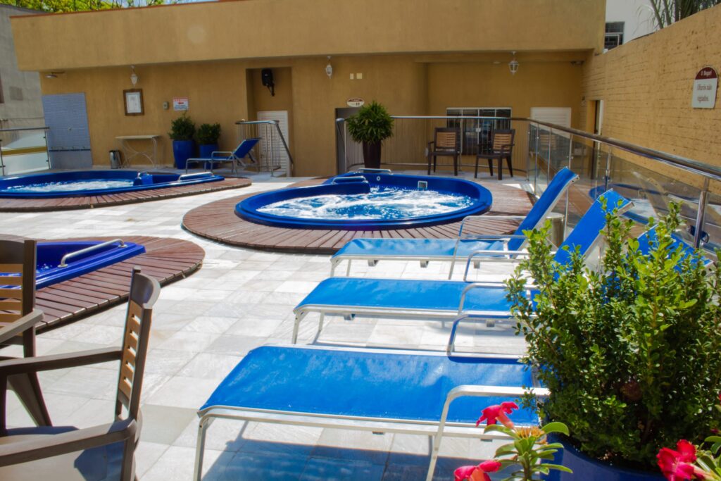 Área de lazer com espreguiçadeiras azuis, banheiras de hidromassagem e decoração com plantas, proporcionando um ambiente relaxante e confortável.