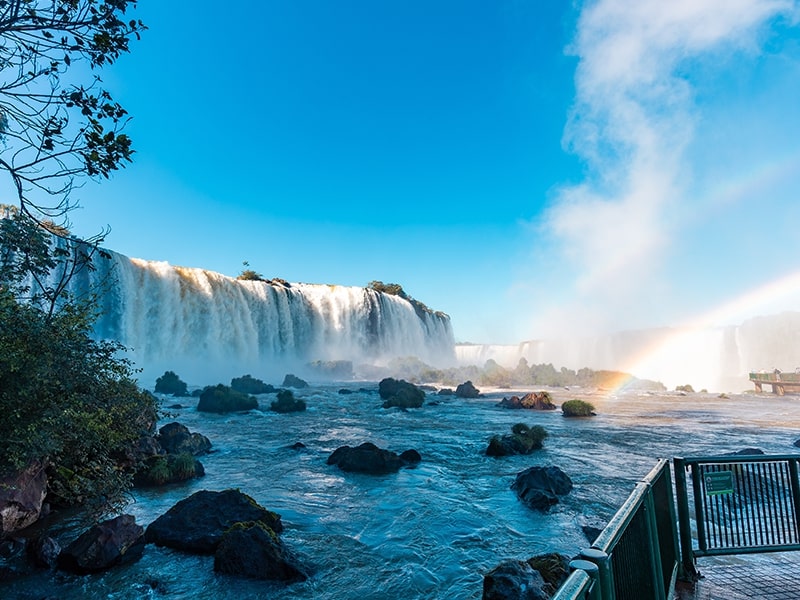 Vista das deslumbrantes Cataratas do Iguaçu em um dia ensolarado, com um arco-íris formado pela névoa das quedas d'água. A paisagem é ideal para quem deseja explorar a natureza em Foz do Iguaçu.